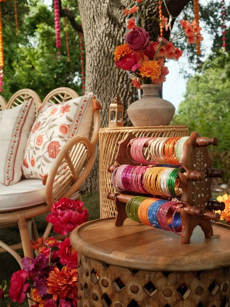 A vibrant outdoor bangle station featuring colorful bangles on a wooden display, surrounded by bright florals, woven seating, and embroidered cushions—perfect for a Mehendi or Sangeet celebration.